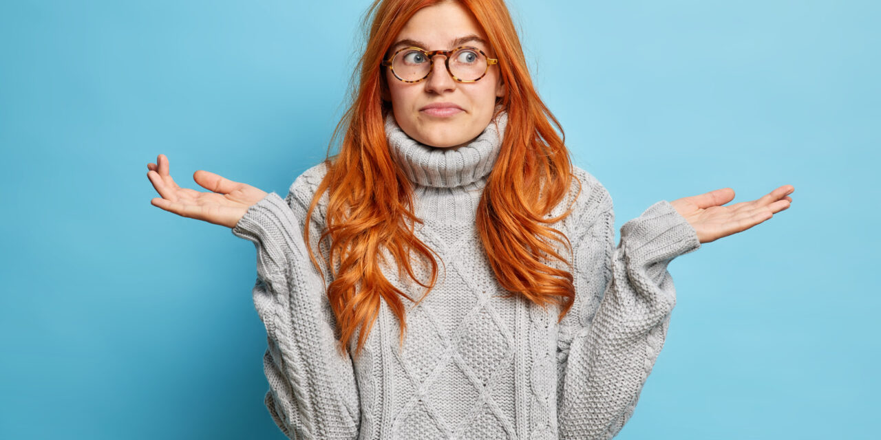 Nice attractive doubtful woman shrugs shoulders doesnt know answer on difficult question doesnt care about your problems stands indifferent against blue wall has no knowledge shows uncertain gesture