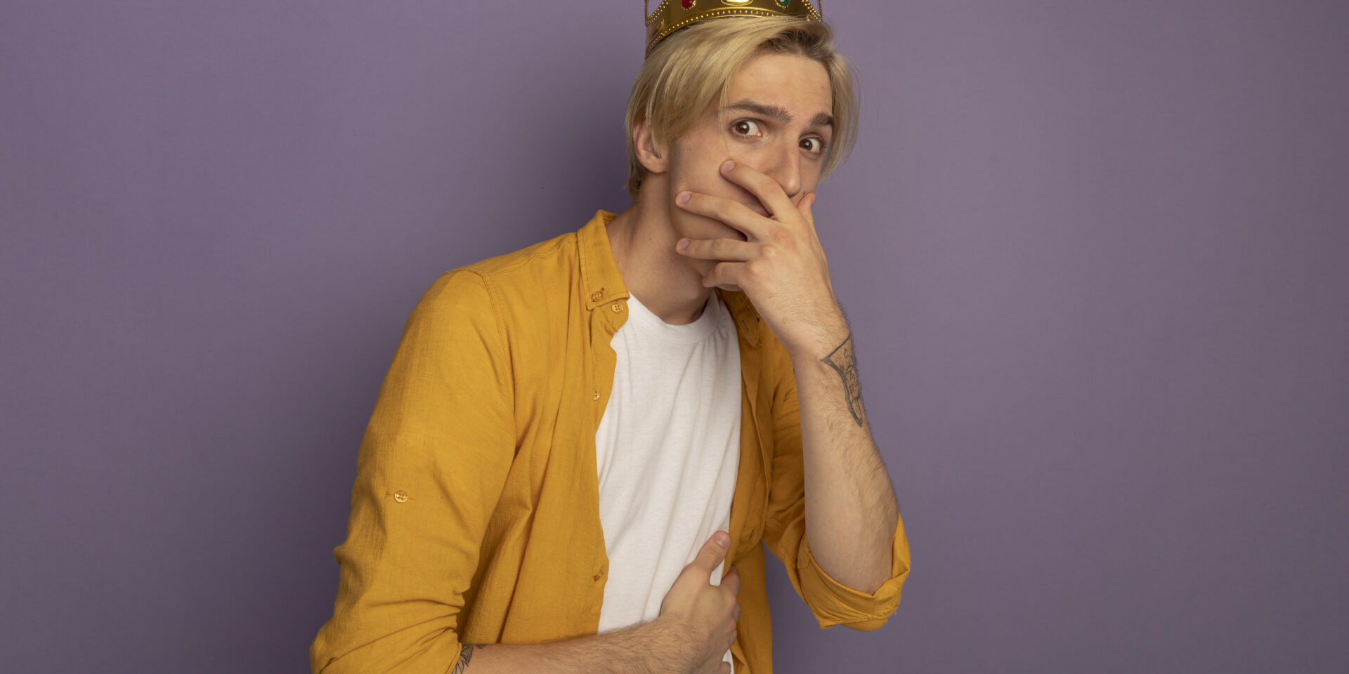 scared young blonde guy wearing yellow t-shirt and crown covered mouth with hand isolated on purple background