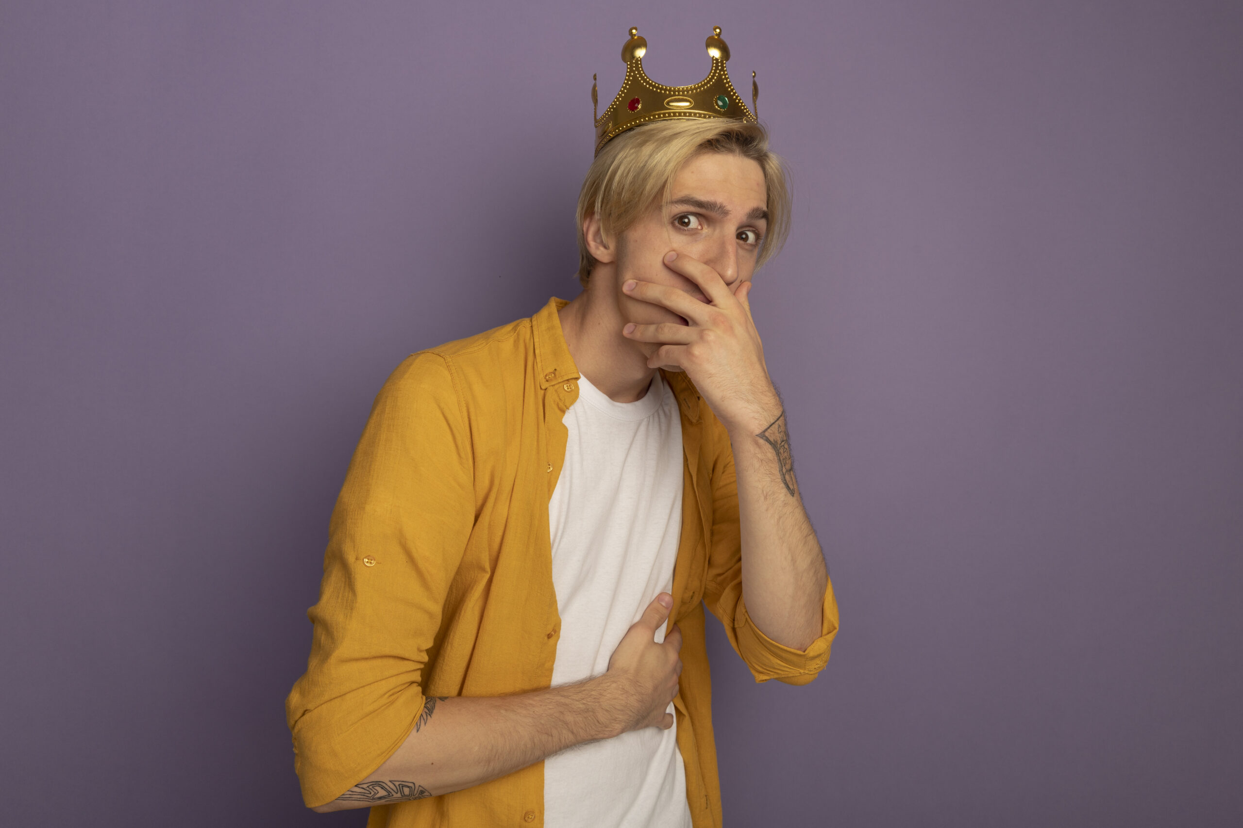 scared young blonde guy wearing yellow t-shirt and crown covered mouth with hand isolated on purple background