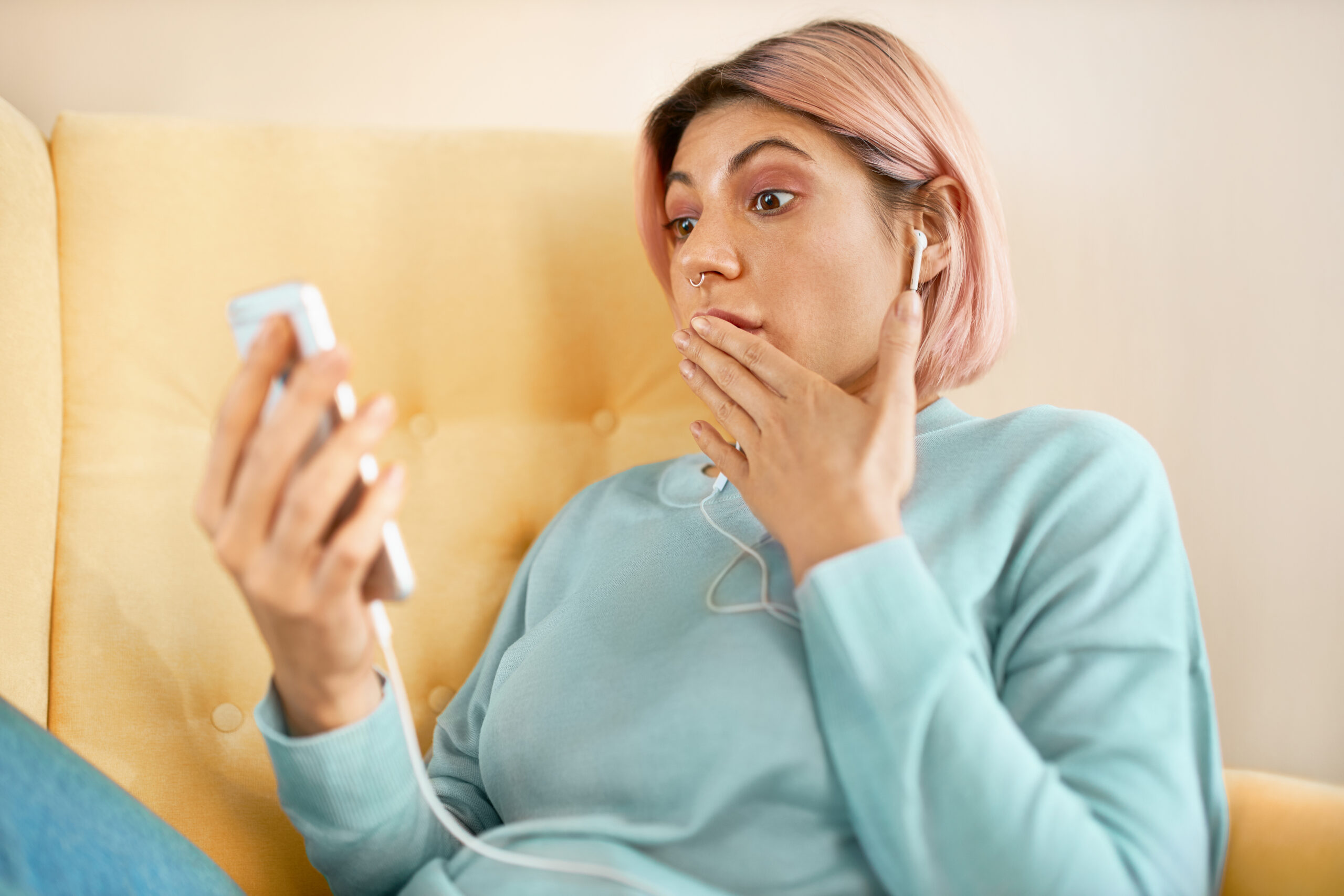 Emotional surprised young woman with pinkish hair sitting on sofa with smart phone, covering mouth in astonishment, gasping, receiving unexpected news while messaging online using home wifi
