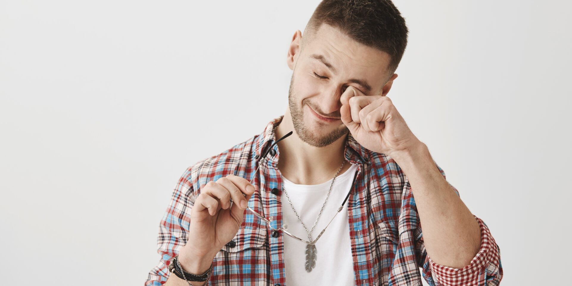 Student tired from working in computer all night. Portrait of sleepy or exhausted handsome male with trendy haircut and outfit, rubbing eyes while holding glasses in hand, standing over gray wall