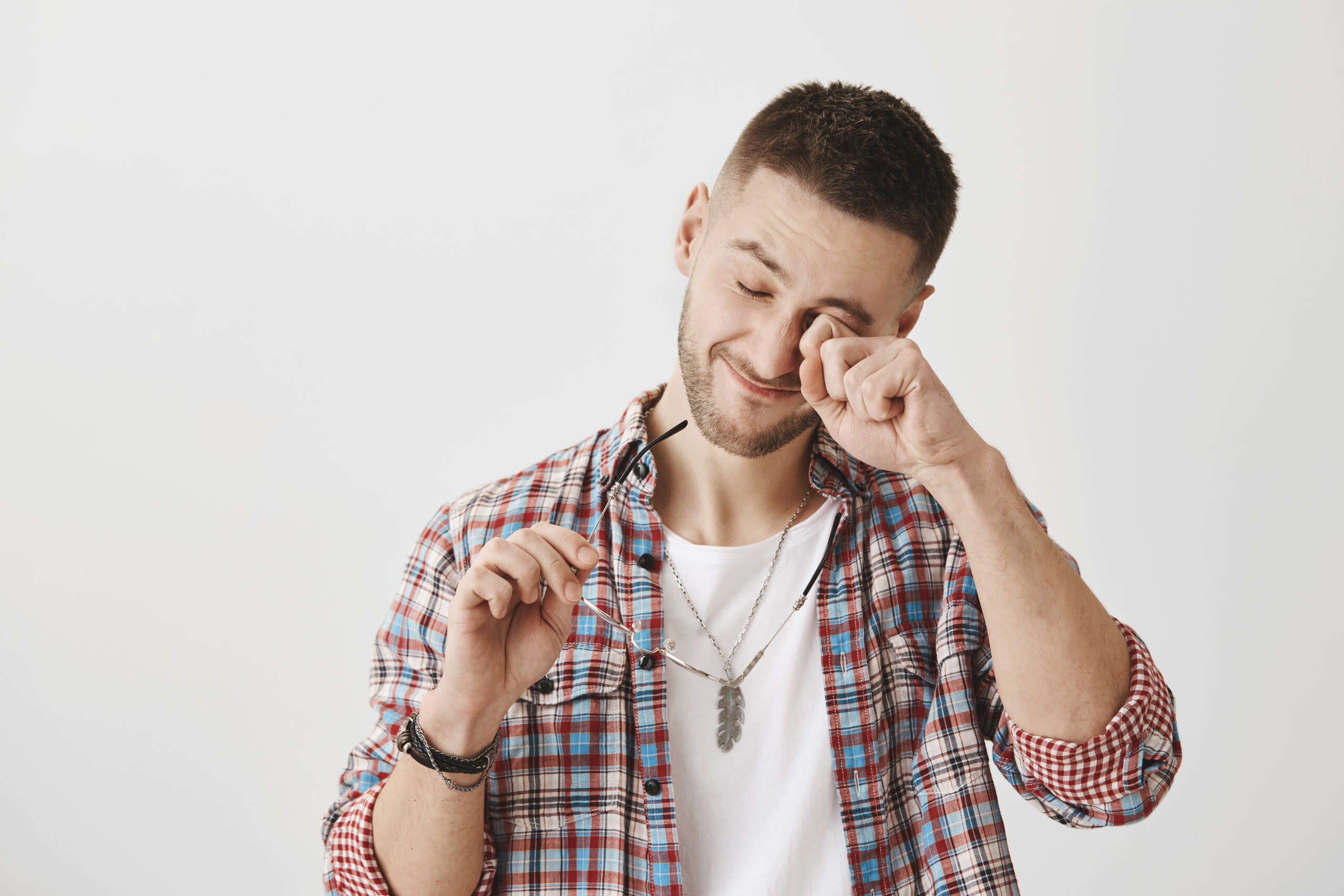 Student tired from working in computer all night. Portrait of sleepy or exhausted handsome male with trendy haircut and outfit, rubbing eyes while holding glasses in hand, standing over gray wall