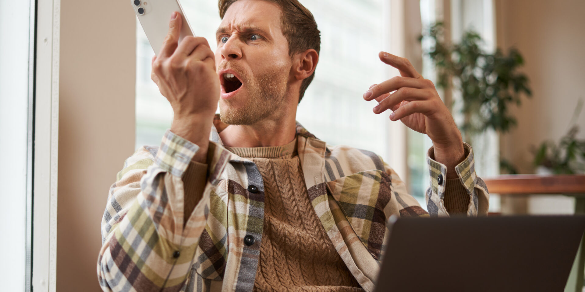 Portrait of an angry man shouting at his mobile phone. Frustrated cafe visitor arguing with someone on call, sitting with laptop near window, screaming at telephone