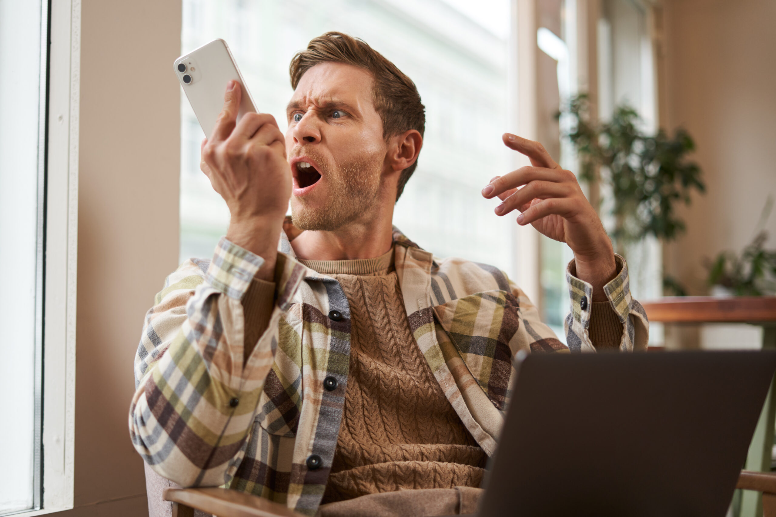Portrait of an angry man shouting at his mobile phone. Frustrated cafe visitor arguing with someone on call, sitting with laptop near window, screaming at telephone
