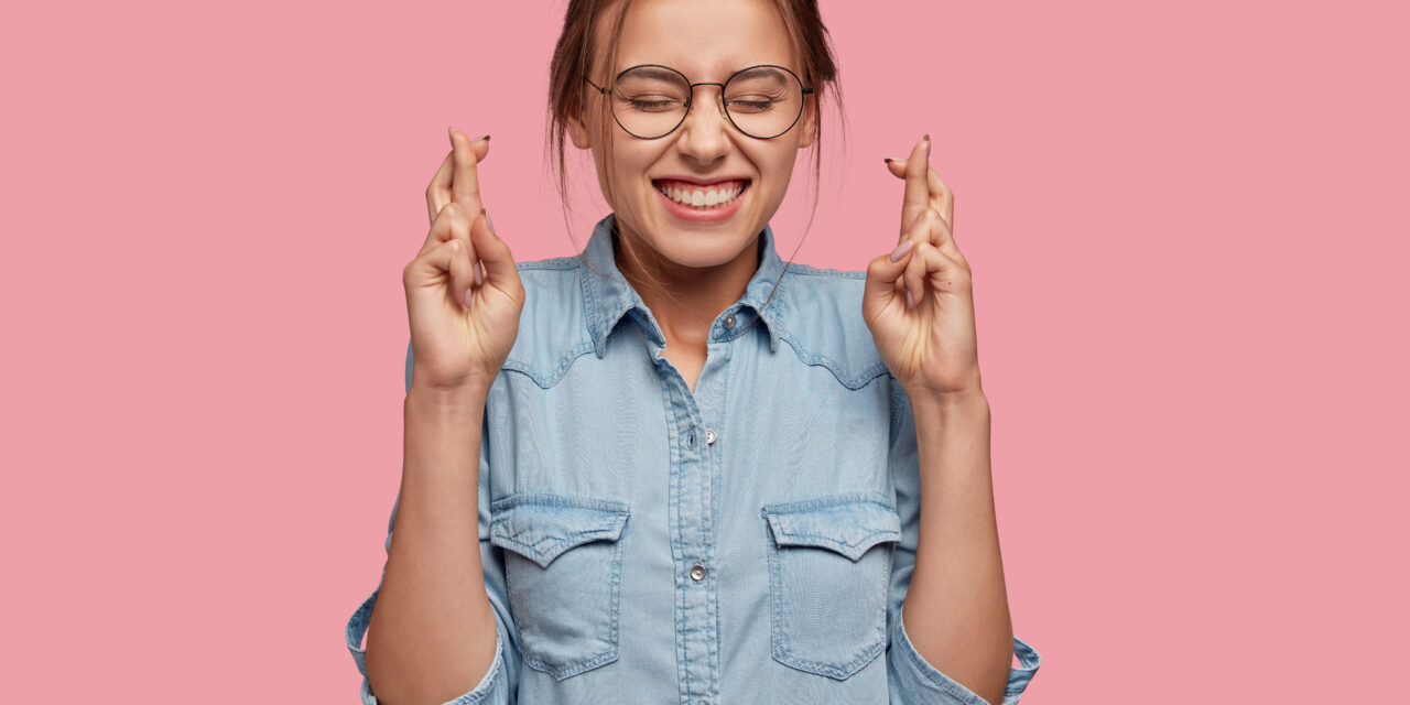 Studio shot of superstitious female with cheerful expression crosses fingers for good luck, believes dreams come true, dressed in stylish denim shirt, isolated over pink background. Wish concept