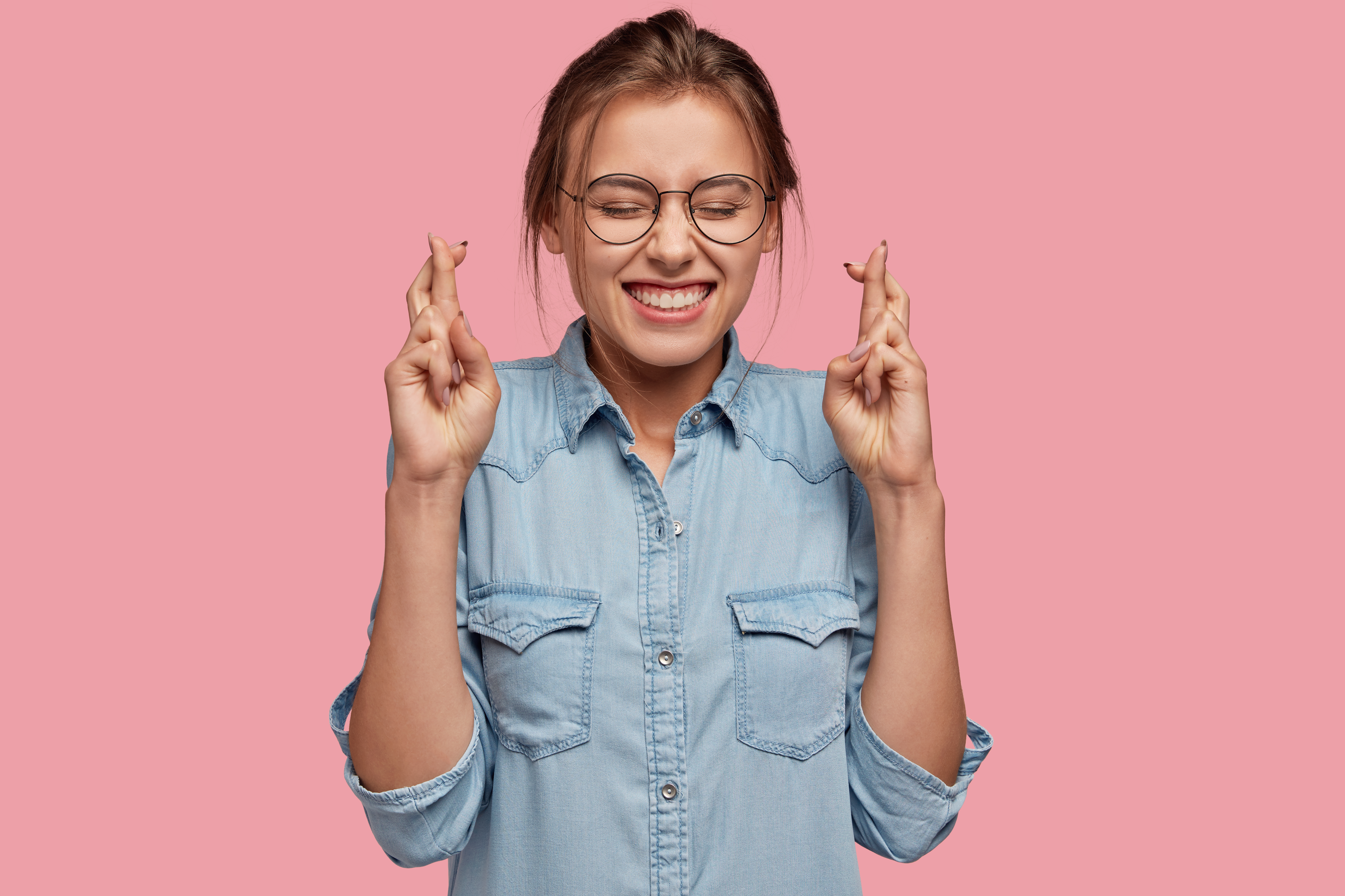 Studio shot of superstitious female with cheerful expression crosses fingers for good luck, believes dreams come true, dressed in stylish denim shirt, isolated over pink background. Wish concept