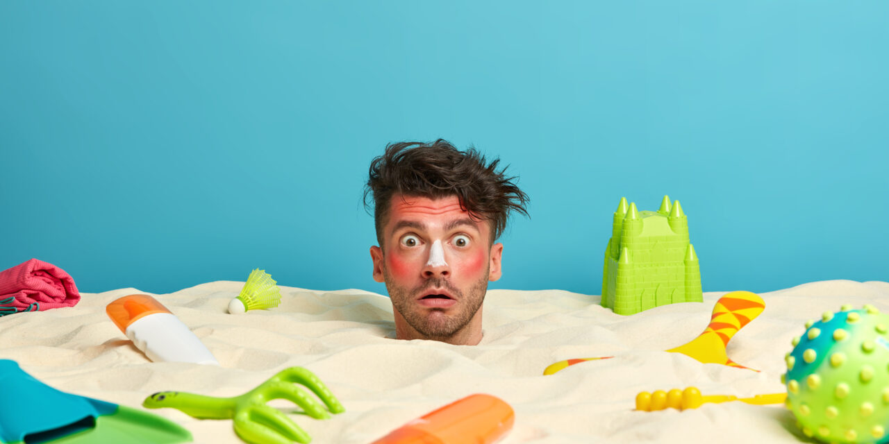 Shocked man with stuck head in sand, red burnt skin, uses sunscreen, spends free time on vacation, surrounded with beach accessories over sea horizon. Summer holidays, recreation, relaxation concept