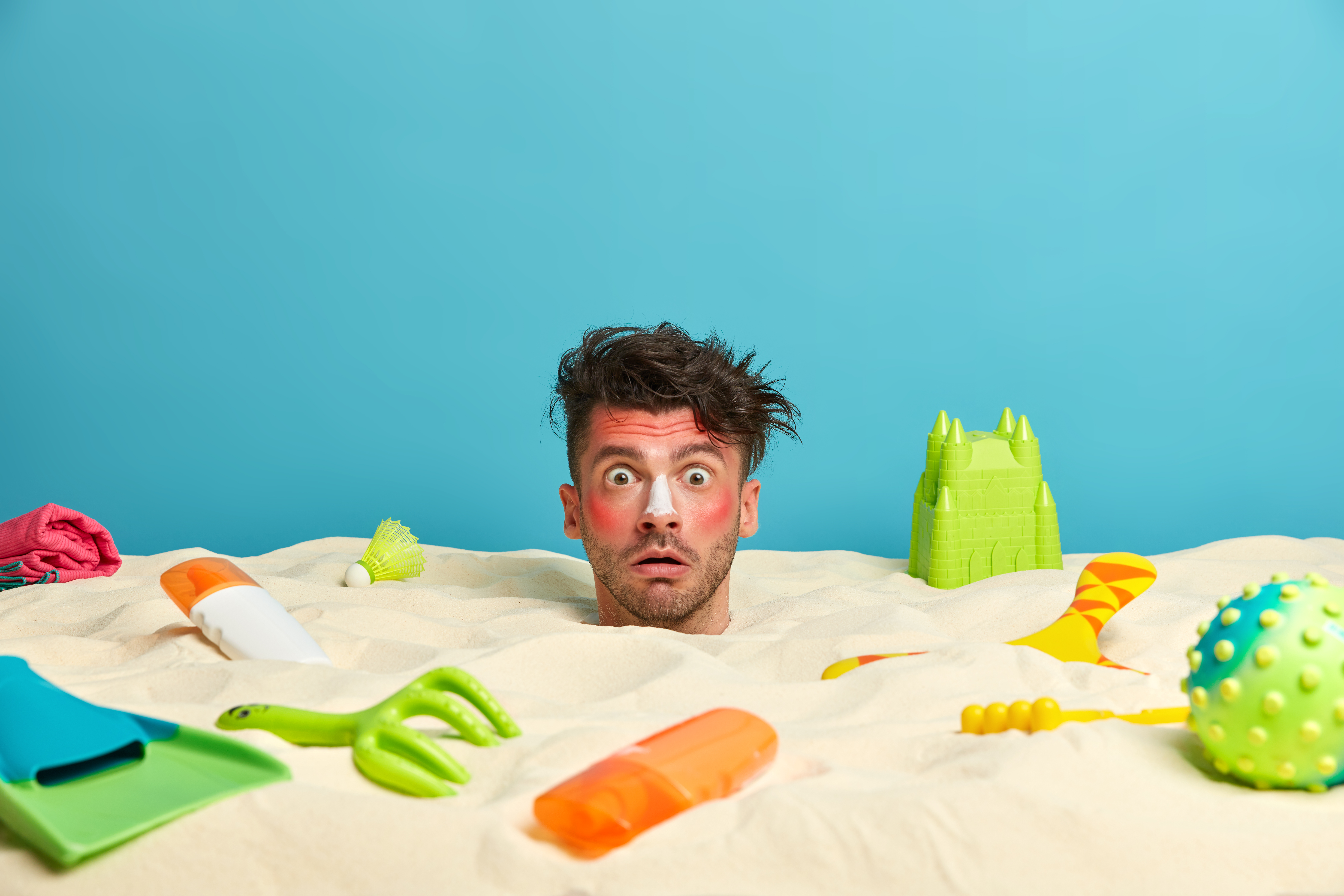 Shocked man with stuck head in sand, red burnt skin, uses sunscreen, spends free time on vacation, surrounded with beach accessories over sea horizon. Summer holidays, recreation, relaxation concept
