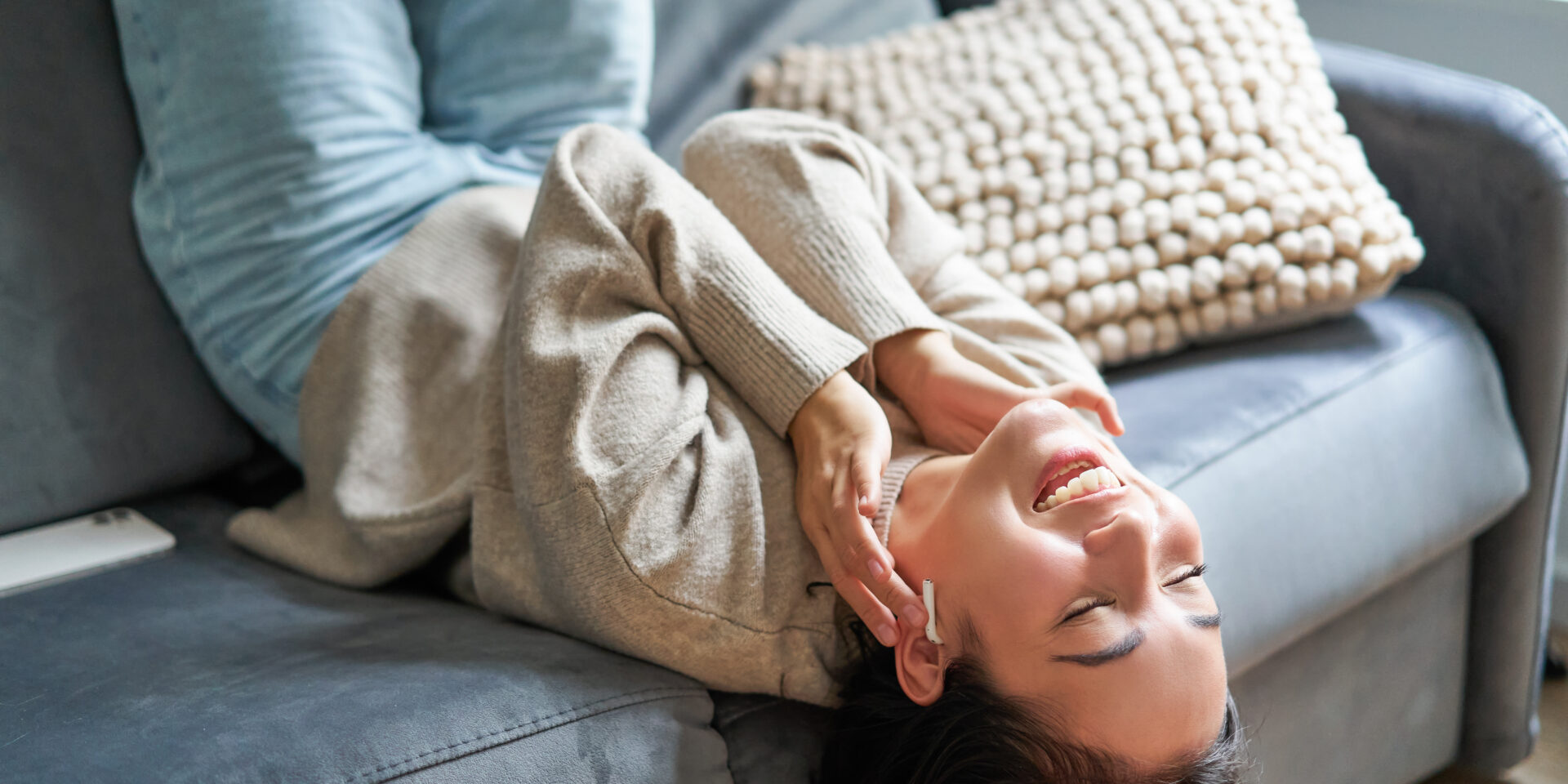 Portrait of happy young asian woman feeling upbeat, lying on sofa and listening music in wireless headphones, smiling pleased