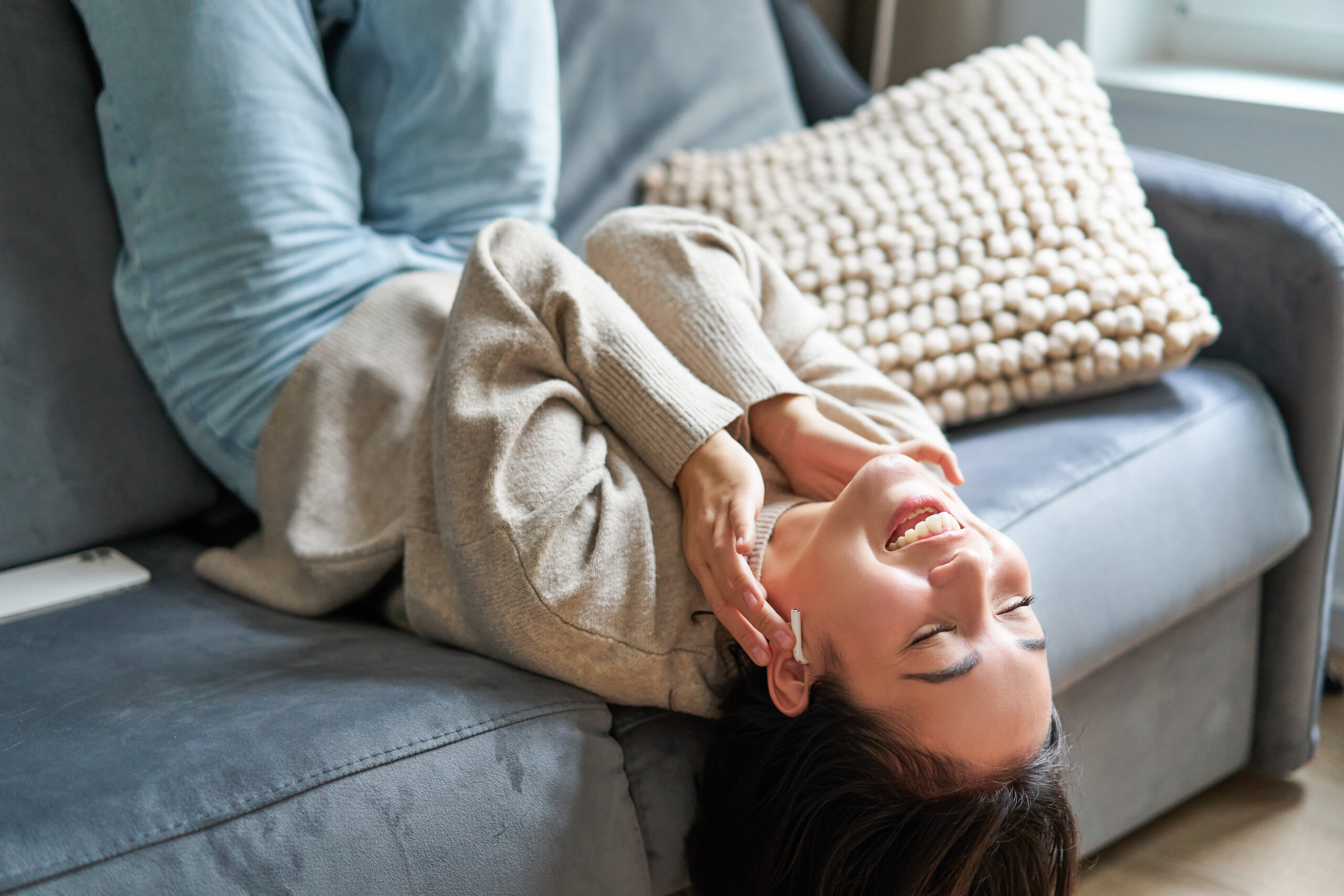 Portrait of happy young asian woman feeling upbeat, lying on sofa and listening music in wireless headphones, smiling pleased