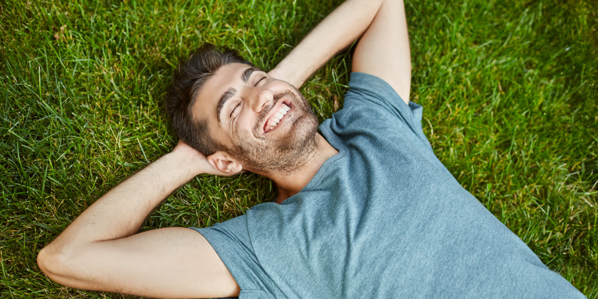 Positive emotions. Young beautiful bearded caucasian male in blue t-shirt lying on grass smiling with teeth, laughing, relaxing outside in summer morning with happy face expression.