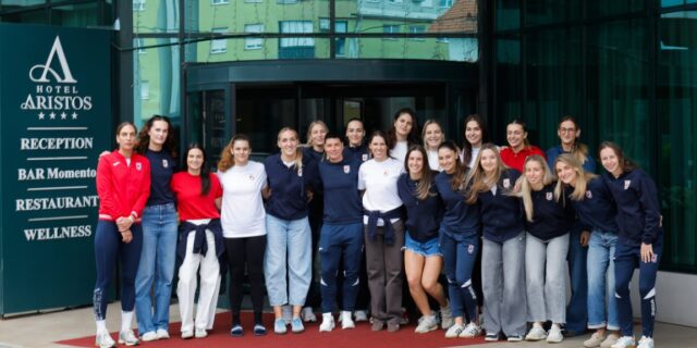 A group of athletes poses together on a red carpet in front of the Hotel Aristos, with a sign listing its amenities.