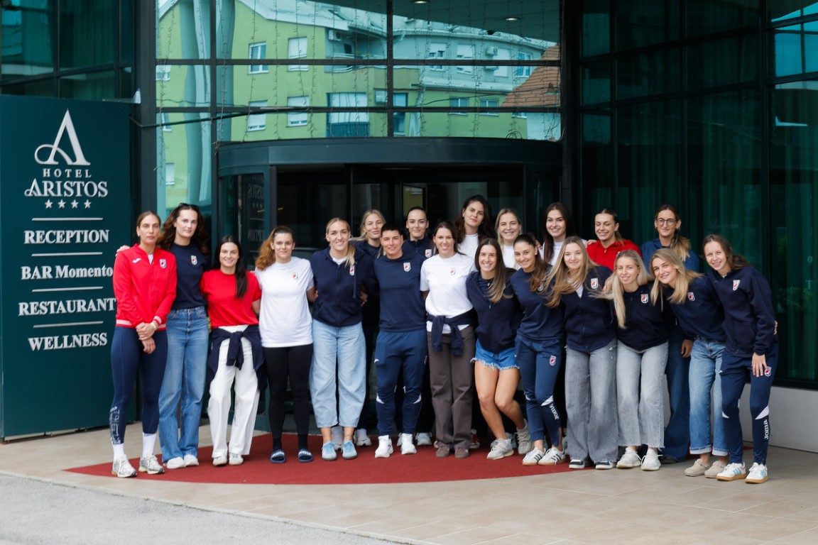 A group of athletes poses together on a red carpet in front of the Hotel Aristos, with a sign listing its amenities.